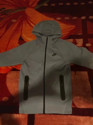 Ljusblå Nike hoodie med dragkedja - Snygg ljusblå hoodie från Nike med hel dragkedja framtill och två svarta dragkedjeförsedda fickor. Klassisk Nike-logga på bröstet och huva för extra stilpoäng. Perfekt för dig som gillar sportig och avslappnad look.