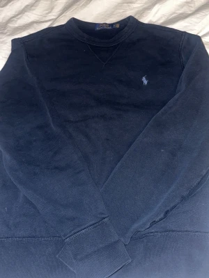 Mörkblå sweatshirt från Polo Ralph Lauren - Mörkblå sweatshirt från Polo Ralph Lauren med klassisk rund halsringning och broderad logotyp på bröstet. Tröjan har långa ärmar och ribbade muddar vid ärmslut och nederkant. Perfekt för en avslappnad och stilren look.
