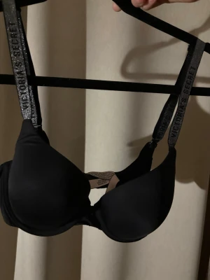 Svart push-up bh från Victoria's Secret - Snygg svart push-up bh från Victoria's Secret med vadderade kupor och justerbara axelband med logotypdetaljer. Bh:n har en slät finish och ger bra stöd och form. Perfekt för dig som vill ha en klassisk och stilren look.
