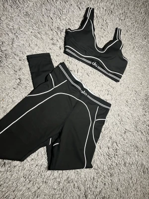 Svart träningsset från Alo - Snyggt svart träningsset från Alo med sport-bh och tights. Setet har vita kontrastsömmar och logga på både bh och midja. Materialet är stretchigt och passar perfekt för gym eller yoga. Sport-bhn har dubbla axelband för extra stöd.