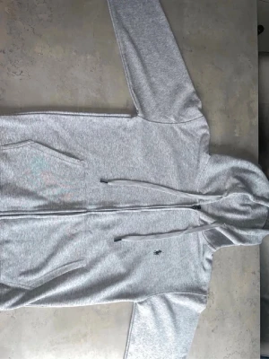 Grå zip-hoodie från Polo Ralph Lauren - Snygg ljusgrå hoodie med dragkedja från Polo Ralph Lauren. Klassisk design med huva och dragsko, två fickor framtill och broderad logga på bröstet. Perfekt för en chill och stilren look. Mjuk bomullsblandning som känns skön mot huden. Hör av er vid frågor! 
