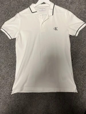 Vit piké från Calvin Klein Jeans - Vit pikétröja från Calvin Klein Jeans med svart CK-logga på bröstet och svarta detaljer på krage och ärmslut. Slim fit-modell med korta ärmar och klassisk krage. Perfekt för en clean och stilren look.
