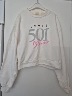 Sweatshirt - Vit croppad sweatshirt från Levi's med stort tryck '501 Blues' i grått och rosa framtill. Tröjan har rund hals, långa ärmar och ribbade muddar. Avslappnad passform, perfekt för en chill look. Materialet ser ut att vara mjuk bomullsblandning. Den har fläckar. Annars är den i bra skick.