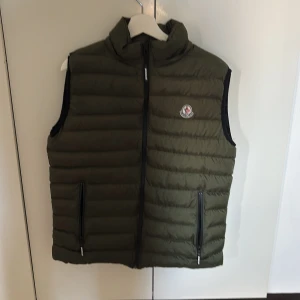 Grön Moncler Väst med luva - Säljer en olivgrön dunväst från Moncler med quiltad design och hög krage. Västen har två fickor med dragkedja och Moncler-logga på bröstet. Insidan har en cool serietidningsetikett. Perfekt för lager-på-lager och streetstyle. Luvan går att ta av.