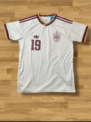 Spanien Lamine Yamal matchtröja - Vit Spanien fotbollströja från Adidas med vinröda detaljer, nummer 19 och Lamine Yamal på ryggen. Tröjan har klassiska Adidas-ränder på axlarna, broderad landslagssköld och guldfärgade inslag vid krage och ärmslut. Perfekt för fotbollsfans!