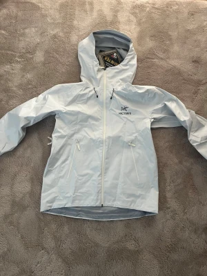 Vit Arc'teryx regnjacka - Snygg vit regnjacka från Arc'teryx med huva och dragkedja framtill. Jackan har två sidofickor med dragkedja och är tillverkad i vattentätt Gore-Tex-material.