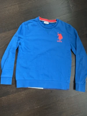 Blå sweatshirt från US Polo Assn - Blå sweatshirt med rund hals från US Polo Assn. Tröjan har röd logga och text på bröstet samt ribbade muddar vid ärmslut och nederkant. Insidan av kragen har en röd detalj som ger en snygg kontrast. Perfekt för en avslappnad och sportig stil. 