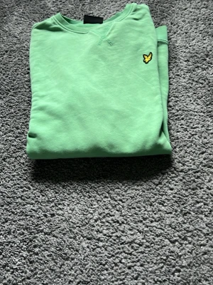 Grön sweatshirt från Lyle & Scott - Snygg grön sweatshirt från Lyle & Scott med klassisk rund hals och det ikoniska gula fågel-loggan broderad på bröstet. Tröjan har långa ärmar och ribbade muddar vid ärmslut och nederkant. Perfekt för en avslappnad och trendig stil.