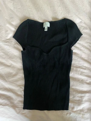 Svart ribbad topp med sweetheart-ringning - Snygg svart ribbad topp med korta ärmar och markerad sweetheart-ringning. Figurnära passform och stretchigt material som ger en modern och trendig look. Perfekt att styla med jeans eller kjol för en cool vibe.