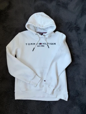 Vit hoodie från Tommy Hilfiger - Snygg vit hoodie från Tommy Hilfiger med broderad logga och text. Klassisk huva med dragsko och ribbade muddar. Materialet är otroligt mjukt och bekvämt, perfekt för en avslappnad stil. Ikonisk flagga broderad på ärmen. Tyvärr som syns på bilderna finns små missfärgningar som jag inte ännu försökt få bort, men som säkerligen enkelt går bort, därav det billiga priset. Skriv vid frågor eller funderingar.