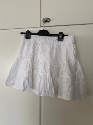Vit volangkjol i bomull - Superfin vit kortkjol i bomull med broderade hål och volangdetaljer. Perfekt för varma dagar och ger en söt och luftig känsla. Resår i midjan för skön passform. Lätt att matcha med både toppar och sneakers.