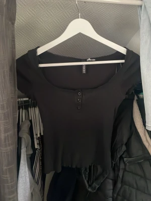 Svart ribbad topp från H&M - Svart ribbad topp från H&M med korta ärmar och rundad halsringning. Toppen har knappar framtill och en lätt croppad passform. Materialet är stretchigt och bekvämt, perfekt för en trendig vardagslook.