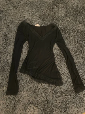 Svart långärmad topp med spets - Svart långärmad topp med v-ringning och fina spetsdetaljer längs halsringningen och nederkanten. Sitter tight och har en feminin look som passar perfekt till jeans eller kjol. Materialet känns mjukt och stretchigt.