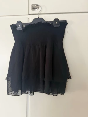 Svart volangig kjol med prickar - Svart kjol med smock upptill och volangig nederdel. Kjolen har ett tunt, transparent lager med små prickar i tyget. Perfekt för en trendig och luftig stil.