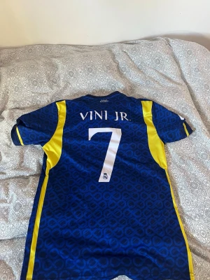 Real Madrid Vini Jr. matchtröja blå - Snygg blå Real Madrid matchtröja från Adidas med gula detaljer och Vini Jr. nummer 7 på ryggen. Tröjan har klubbmärke och sponsortryck på bröstet samt klassiska Adidas-ränder på axlarna. Perfekt för fotbollsfans som vill sticka ut på planen eller läktaren.