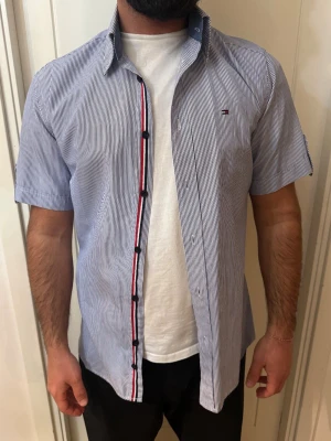 Randig kortärmad skjorta Tommy Hilfiger -  blå- och vitrandig skjorta från Tommy Hilfiger med korta ärmar och klassisk krage. Skjortan har knappar framtill och diskret logga broderad på bröstet. Perfekt för en stilren look.