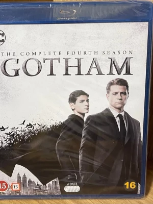 Gotham Säsong 4 Blu-ray box - Gotham The Complete Fourth Season på Blu-ray med fyra skivor. Omslaget är svartvitt med två huvudkaraktärer i kostym och stadssilhuett i bakgrunden. Serien handlar om Gotham Citys brottslighet och kända karaktärer från Batman-universumet.
