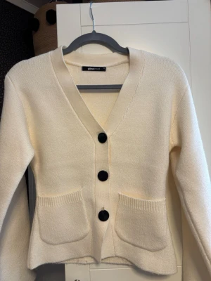 Krämvit kofta från Gina Tricot - Stilren krämvit kofta från Gina Tricot med tre stora svarta knappar framtill och två rymliga fickor. Koftan har v-ringning, lång ärm och är stickad i ett mjukt material. Jätte fint skick, knappt använd.