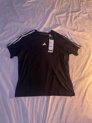 Svart Adidas t-shirt med vita ränder - Svart t-shirt från Adidas med klassiska vita ränder längs axlarna och ett litet Adidas-logotyp på bröstet. T-shirten har rund hals och är kortärmad, perfekt för en sportig och avslappnad stil. Helt ny med prislapp. Fick i present men var för liten. Köptes för 250 kr. 