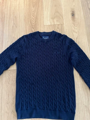 Mörkblå kabelstickad tröja Bondelid - Marinblå kabelstickad tröja från Bondelid i storlek S med rund halsringning och ribbade muddar. Klassisk design med tydlig struktur i stickningen och diskret broderad logga på bröstet. Tillverkad i 100% bomull. Perfekt för dig som gillar stilrena och tidlösa plagg.