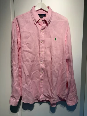 Rosa linneskjorta från Ralph Lauren - Snygg rosa skjorta från Ralph Lauren i linne med klassisk krage och knäppning framtill. Skjortan har lång ärm och den ikoniska gröna loggan broderad på bröstet. Perfekt för en avslappnad look nu till sommaren. Storlek XL!