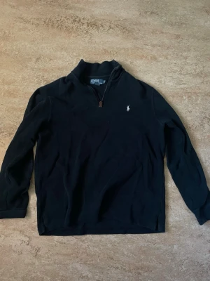 Mörkblå tröja från Polo Ralph Lauren - Mörkblå långärmad tröja från Polo Ralph Lauren i storlek M med halv dragkedja och klassisk broderad logga på bröstet. Tröjan har en hög krage och är tillverkad i mjukt material som passar perfekt till en avslappnad stil. 100% bomull.