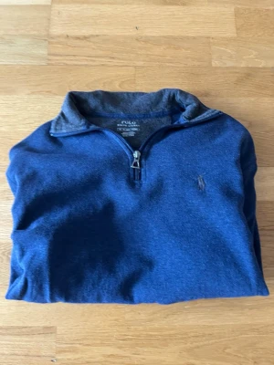 Blå halvzip tröja från Polo Ralph Lauren - Mörkblå tröja med halv dragkedja från Polo Ralph Lauren. Klassisk passform med broderad logga på bröstet och ribbade muddar. Perfekt för en avslappnad men ändå stilren look.