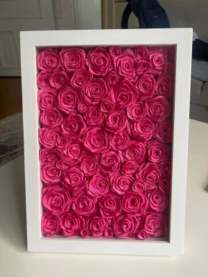 Tavla med rosa konstgjorda rosor - En dekorativ tavla med massor av tätt placerade konstgjorda rosor i stark rosa färg, inramad i en vit, rektangulär träram. Perfekt som färgglad inredningsdetalj till rummet eller som present.