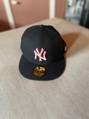 Svart New Era Yankees keps rosa logo - Snygg svart New Era 59FIFTY keps med klassisk New York Yankees-logga i rosa framtill. Kepsen har rak skärm och broderade detaljer i rosa även på sidan och bak. Perfekt för dig som vill sticka ut med en sportig och trendig look.