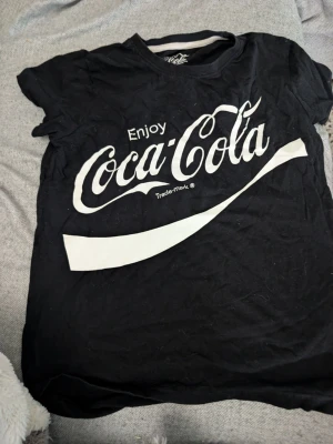 Svart Coca-Cola t-shirt stl 152 - Cool svart t-shirt från Coca-Cola med stort vitt tryck på framsidan. Klassisk rund hals och korta ärmar. Perfekt för dig som gillar retro och streetwear vibes.