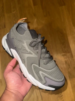 Louis Vuitton grå tatic sneakers - Snygga grå sneakers från Louis Vuitton med stickad ovandel, gråa och vita detaljer samt robust sula. Skorna har en modern design, LV-logga på sidan och läderdetalj i brunt på hälen. Perfekta för dig som gillar exklusiva och trendiga sneakers.