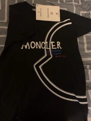 Svart Moncler t-shirt med tryck - Svart t-shirt från Moncler med stort vitt och blått tryck framtill och logga. Klassisk passform med korta ärmar och rund hals. Snygg kontrastdetalj i vitt som går diagonalt över framsidan. Perfekt för dig som gillar streetwear och exklusiva märken.