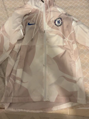 Chelsea FC vindjacka från Nike - Snygg vindjacka från Nike med Chelsea FC-logga på bröstet. Jackan har ett abstrakt mönster i beige och vitt, hel dragkedja framtill och huva med dragsko. Perfekt för dig som vill visa ditt stöd för Chelsea och samtidigt hålla stilen.
