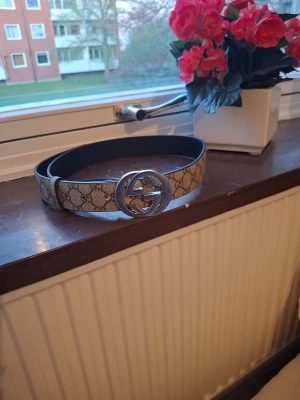 Gucci beige bälte med GG-spänne - Snyggt beige bälte från Gucci med klassiskt GG-mönster och ett stort, rundat silverspänne med GG-logga. Bältet har en exklusiv känsla och passar perfekt till jeans eller kostymbyxor.