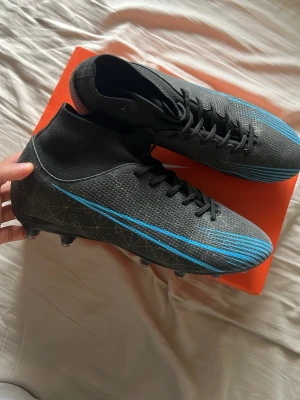 svarta fotbollsskor med blå detaljer - Säljer ett par svart fotbollsskor med blåa linjer och diskret mönster på sidorna. Skorna har högt skaft, snörning och är tillverkade i syntetmaterial för bra grepp och kontroll på planen. Perfekta för dig som vill sticka ut på planen. (OBS det är inte nike)