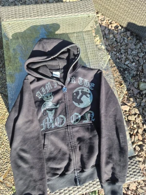 Aim for the moon - Svart hoodie med dragkedja och huva från Urban Classics. Aim for the moon  Framsidan har ett stort tryck med texten 'AIM FOR THE MOON', en tecknad figur med krona och en måne i glittrande stenar. Perfekt för dig som gillar streetwear och unika detaljer.