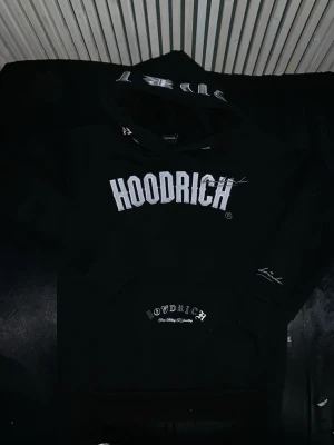 Äkta Hoodrich Hoodie - Svart hoodie från Hoodrich med stor vit logga framtill och tryck på både huva och ficka. Baksidan har texttryck i vitt. Klassisk känguruficka och huva. Perfekt för streetwear-stilen. Vid frågor eller bud är det bara att kontakta mig. Den är såklart äkta!