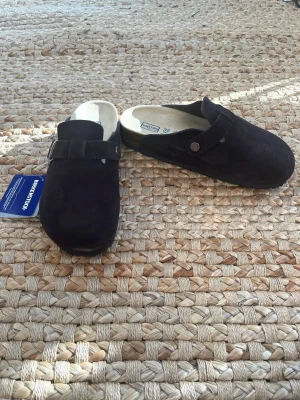 Birkenstock Boston - Säljer ett par brun Birkenstock Boston tofflor i mocka med mjuk fotbädd. Klassisk slip-in modell med justerbart spänne och bekväm innersula. Perfekta för dig som gillar stilrena och sköna skor. 