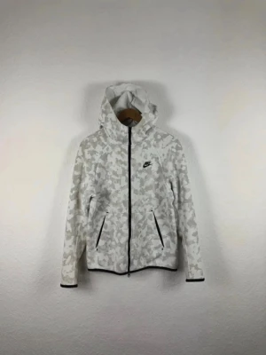 Nike hoodie med beige camomönster - Nike hoodie i vitt och beige camomönster med svart dragkedja och svarta detaljer. Hoodien har två fickor framtill och en justerbar huva. Tillverkad i mjukt och bekvämt material, perfekt för en avslappnad streetwear-look.