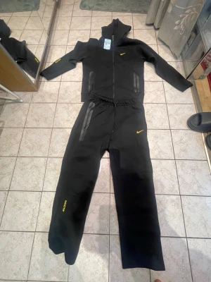 Nike NOCTA svart  - Nike NOCTA svart träningsset med huva och dragkedja. Setet har gula detaljer, inklusive Nike-logga och NOCTA-tryck. Jackan har två dragkedjefickor och byxorna har resår i midjan samt dragsko. Perfekt för sport eller chill. Materialet är mjukt och stretchigt.