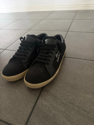 Svarta Saint Laurent sneakers - Svarta sneakers från Saint Laurent med vit logga på sidan. Skorna har svarta skosnören, svart ovandel i mocka och läder samt vit sula. Klassisk låg modell med stilren design, perfekt för dig som gillar exklusiva streetwear-vibbar.