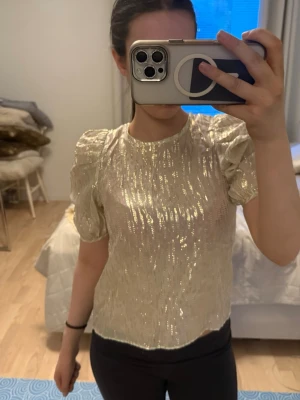 Glittrig guldig topp med puffärm - Snygg glittrig topp i guld med korta puffärmar och rund halsringning. Toppen har en rak passform och skimrande yta som fångar ljuset. Perfekt för dig som vill sticka ut och addera lite extra glam till din outfit.