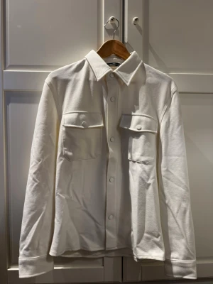 Vit overshirt med fickor och knappar - Stilren vit overshirt med klassisk krage och två bröstfickor med lock och knappar. Overshirten har långa ärmar och knäppning framtill. Perfekt för en clean och modern look.