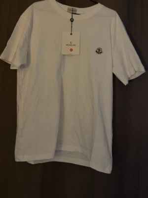 Vit Moncler t-shirt med logga - Snygg vit t-shirt från Moncler med klassisk rund halsringning och korta ärmar. På bröstet sitter Monclers ikoniska logotyp. T-shirten är i mjuk bomull och har en clean, stilren look som passar perfekt till jeans eller shorts.