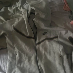 Grå NikeTech - Grå Nike tech tröja men ganska många tecken på användning. Dragkedjan funkar inte men den går att laga, loggan är hel. Den är i storlek XS men skulle säga att den mer sitter som en S