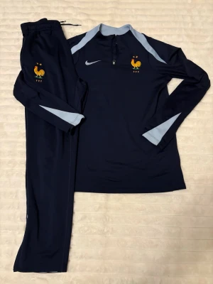 Frankrike Nike träningsset marinblå - Snyggt marinblått träningsset från Nike med Frankrikes fotbollsförbunds logga på både tröja och byxa. Setet har ljusblå detaljer på ärmar och axlar samt dragkedja vid halsen. Perfekt för fotbollsträning eller sportiga dagar.