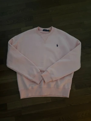 Ljusrosa sweatshirt från Polo Ralph Lauren - Ljusrosa sweatshirt från Polo Ralph Lauren med klassisk rund halsringning och broderad logga på bröstet. Tröjan har långa ärmar, ribbade muddar och är perfekt för en avslappnad stil.