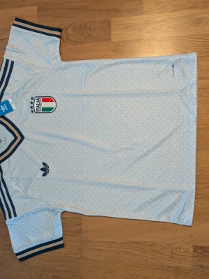 Italien nya fotbollströja Adidas 2026. S.m storlek - Snygg ljusblå nu fotbollströja från Adidas med Italiens landslagsemblem på bröstet och mörkblå detaljer på krage och ärmslut. Tröjan har ett diskret mönster över hela tyget och klassisk v-ringning. Perfekt för dig som gillar fotbolls sportstil.