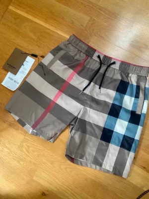 Burberry shorts  - Snygga shorts från Burberry med klassiskt rutigt mönster i beige, svart, blått och rött. Shortsen har elastisk midja med svart snörning och är tillverkade i ett lätt och bekvämt material. Perfekta för sommaren och ger en trendig look.
