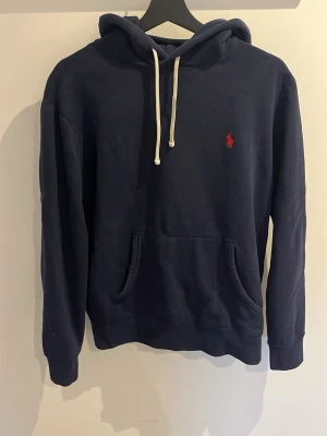 Mörkblå hoodie från Ralph Lauren - Mörkblå hoodie från Ralph Lauren med klassisk röd broderad logga på bröstet. Hoodien har vit dragsko, känguruficka och mjukt material som känns skönt mot huden. Perfekt för en avslappnad och stilren look. Kan gå ner i pris vid snabb affär🙌